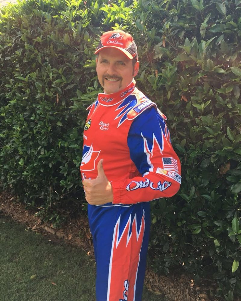 Zac Perkins in a full red and blue NASCAR-style racing suit and cap with a handlebar mustache, giving a thumbs up in front of a hedge